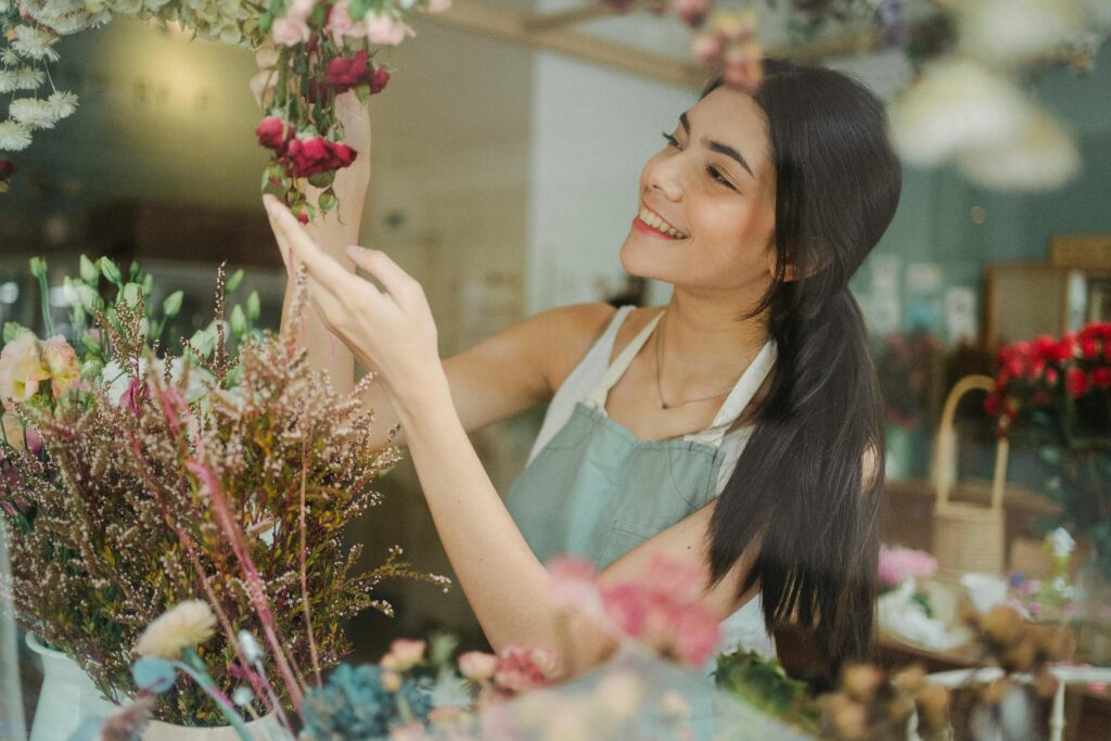 Young smiling dark haired female florist in apron arranging flowers while working in floral shop