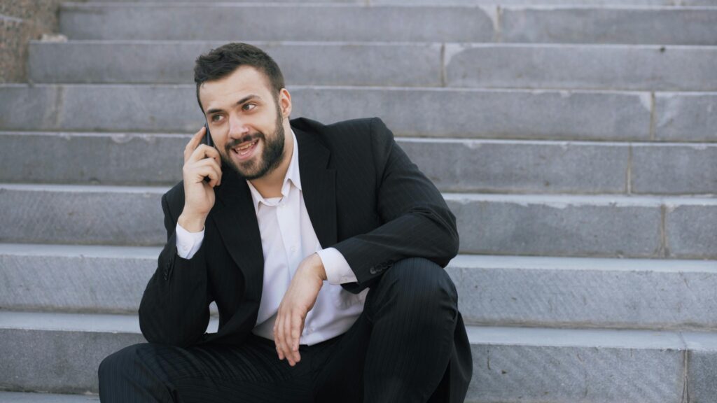 Confident businessman in a suit talking on a phone while sitting on outdoor steps, showcasing modern communication.