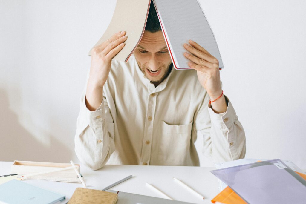 A stressed adult male worker sits at a desk with open notebooks, exhibiting signs of frustration and burnout.