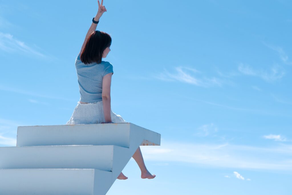 A woman sits on white stairs with blue sky background, reaching upward, embracing freedom.