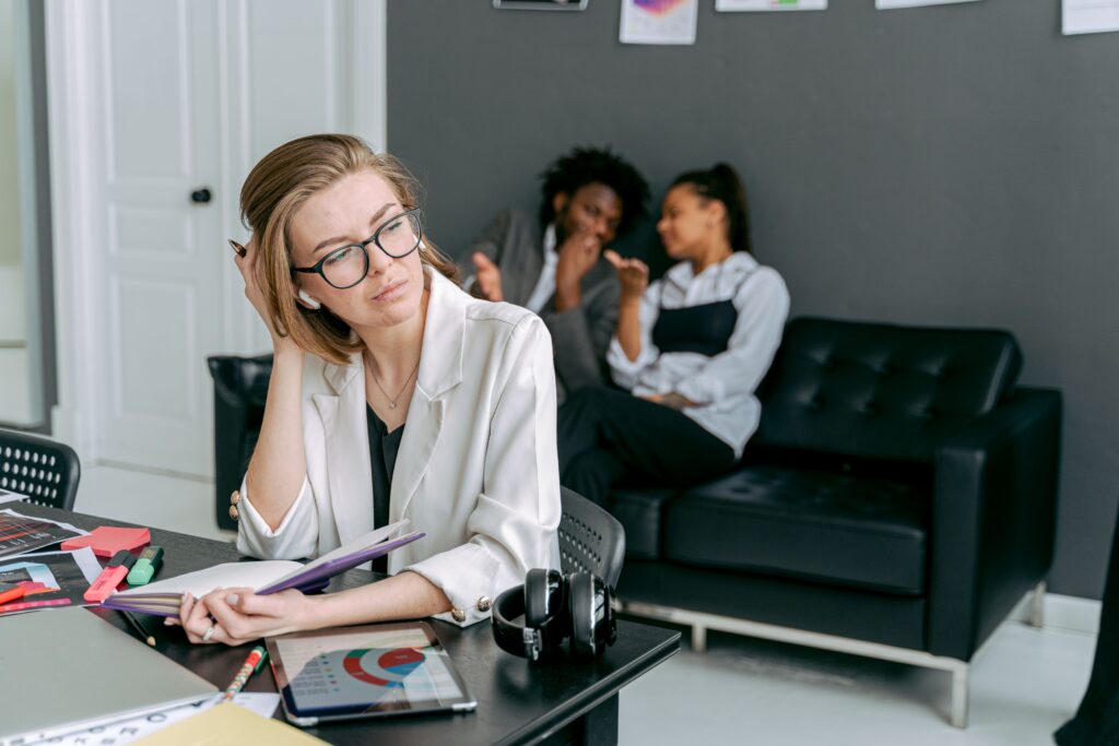 A woman appears stressed at her desk, with colleagues gossiping in the background.