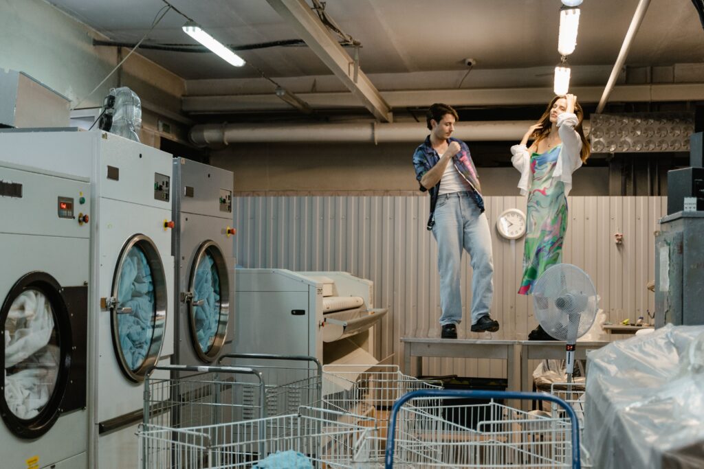 Two adults dancing in an industrial laundry with large machines and carts visible.