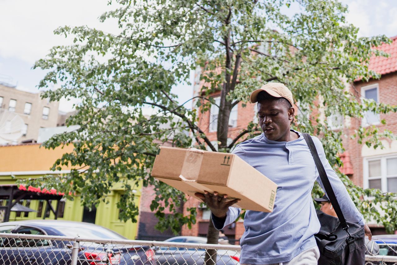A delivery man walks through an urban neighborhood carrying a cardboard package amidst residential buildings.