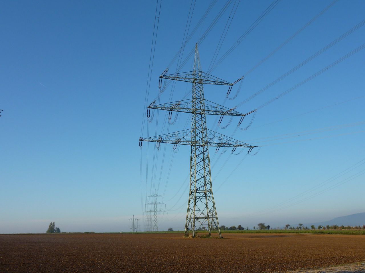 High voltage power lines on a tower standing in an open field against a clear blue sky.