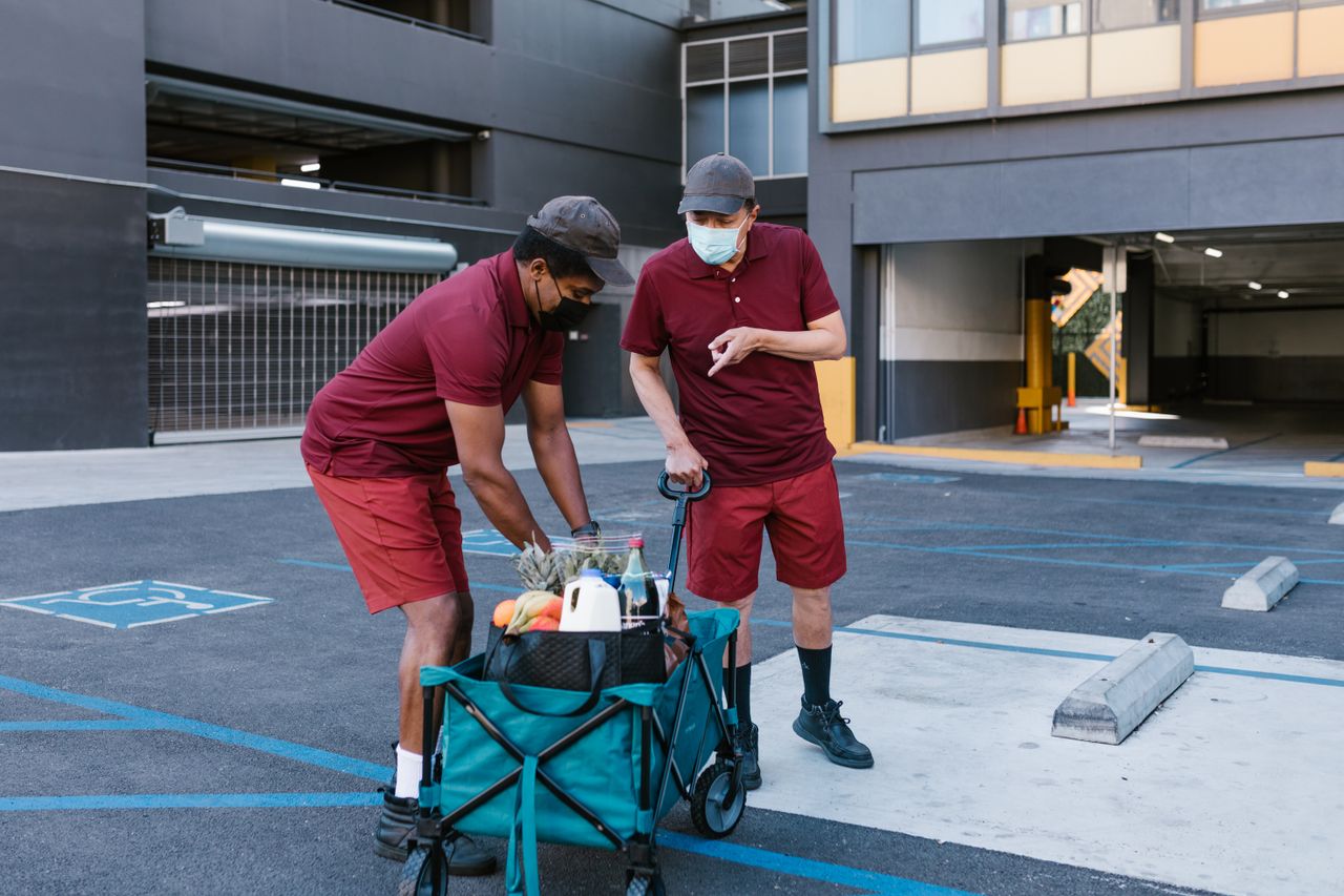 Two masked men in maroon shirts perform food delivery in a parking area with a cart.