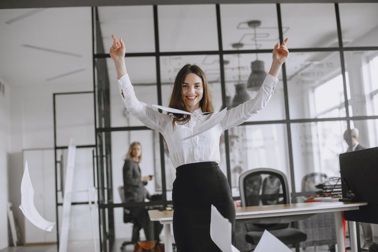 Smiling businesswoman in a modern office throws papers in the air, expressing joy and success.