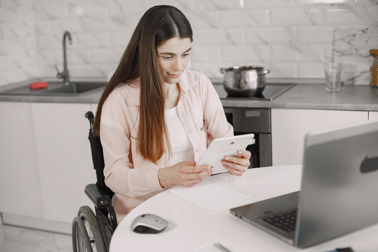 Woman in wheelchair engaged with tablet at kitchen table, creating an inclusive work environment.