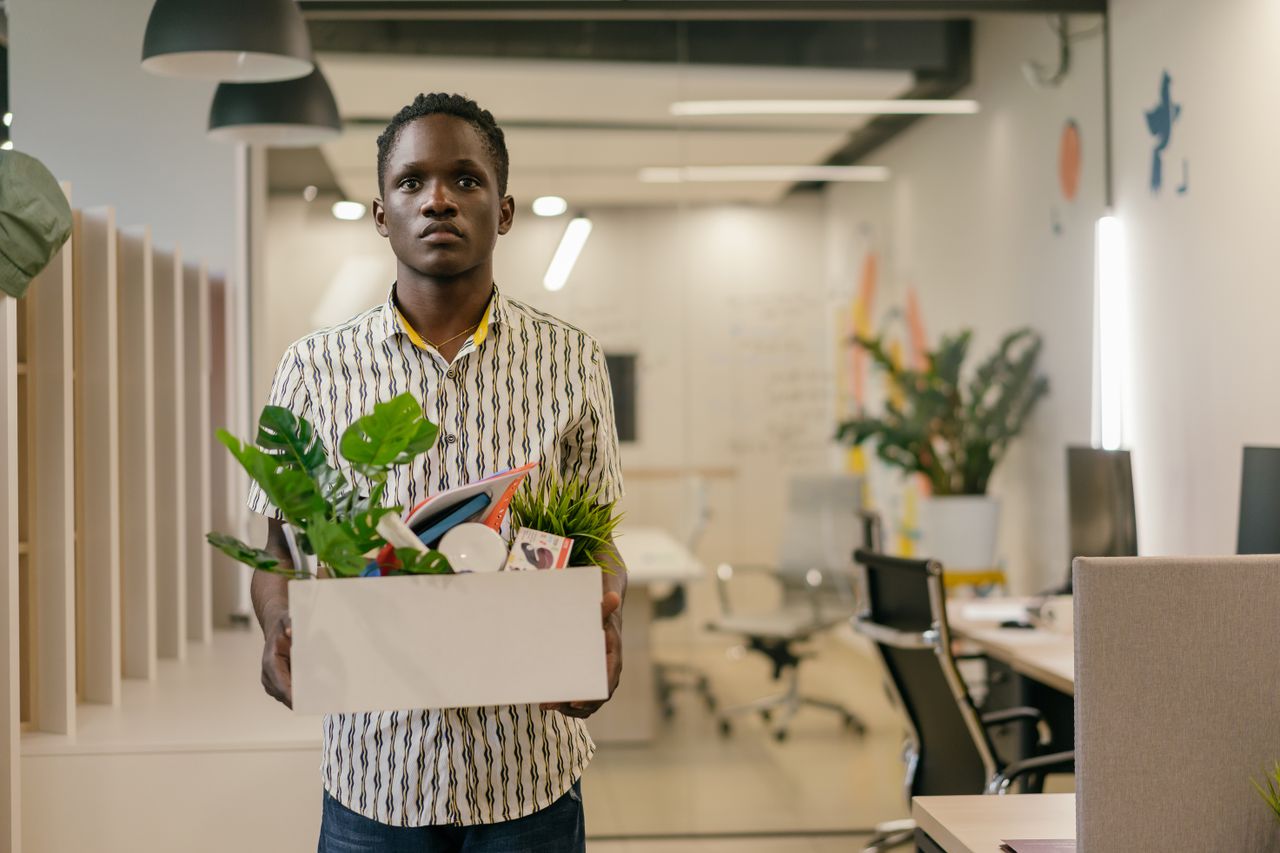 A serious man carries a box of belongings in an office setting, symbolizing dismissal or job change.