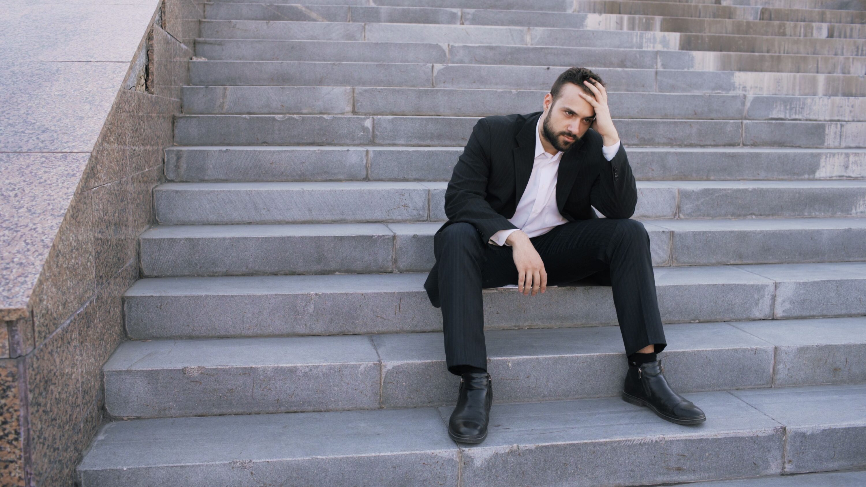 A businessman in a suit sits on stone steps, looking stressed and thoughtful outdoors.
